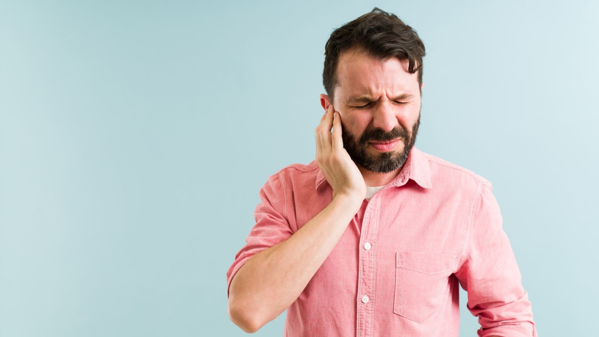 Hombre en camisa salmón, ojos cerrados y haciendo una mueca, llevando la mano a su oído izquierdo sobre fondo azul pálido, que ilustra la molestia característica sentida durante un acúfeno o una fatiga auditiva
