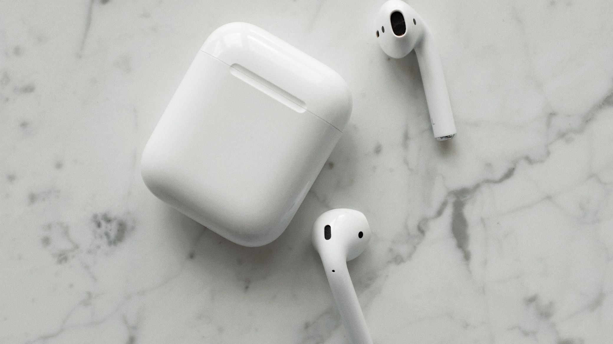 Boîtier d'écouteurs sans fil AirPods classiques en plan flat-lay sur fond marbré blanc, écouteurs semi-in-ear posés à côté, illustrant le format semi-ouvert sans embout silicone