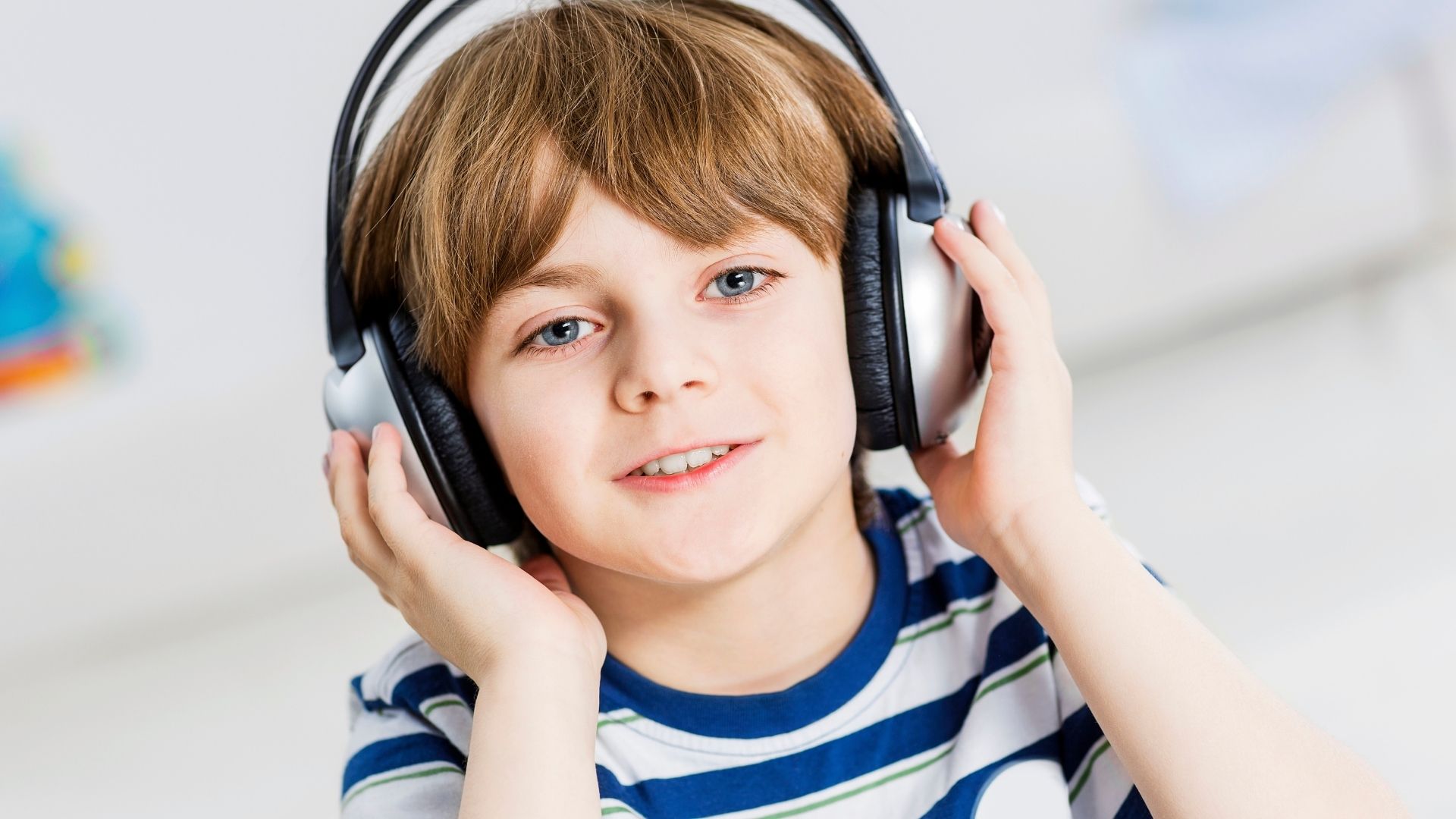 Kind hält mit beiden Händen einen silbernen und schwarzen circum-aural Kopfhörer, ruhiger Gesichtsausdruck, heller Innenraum