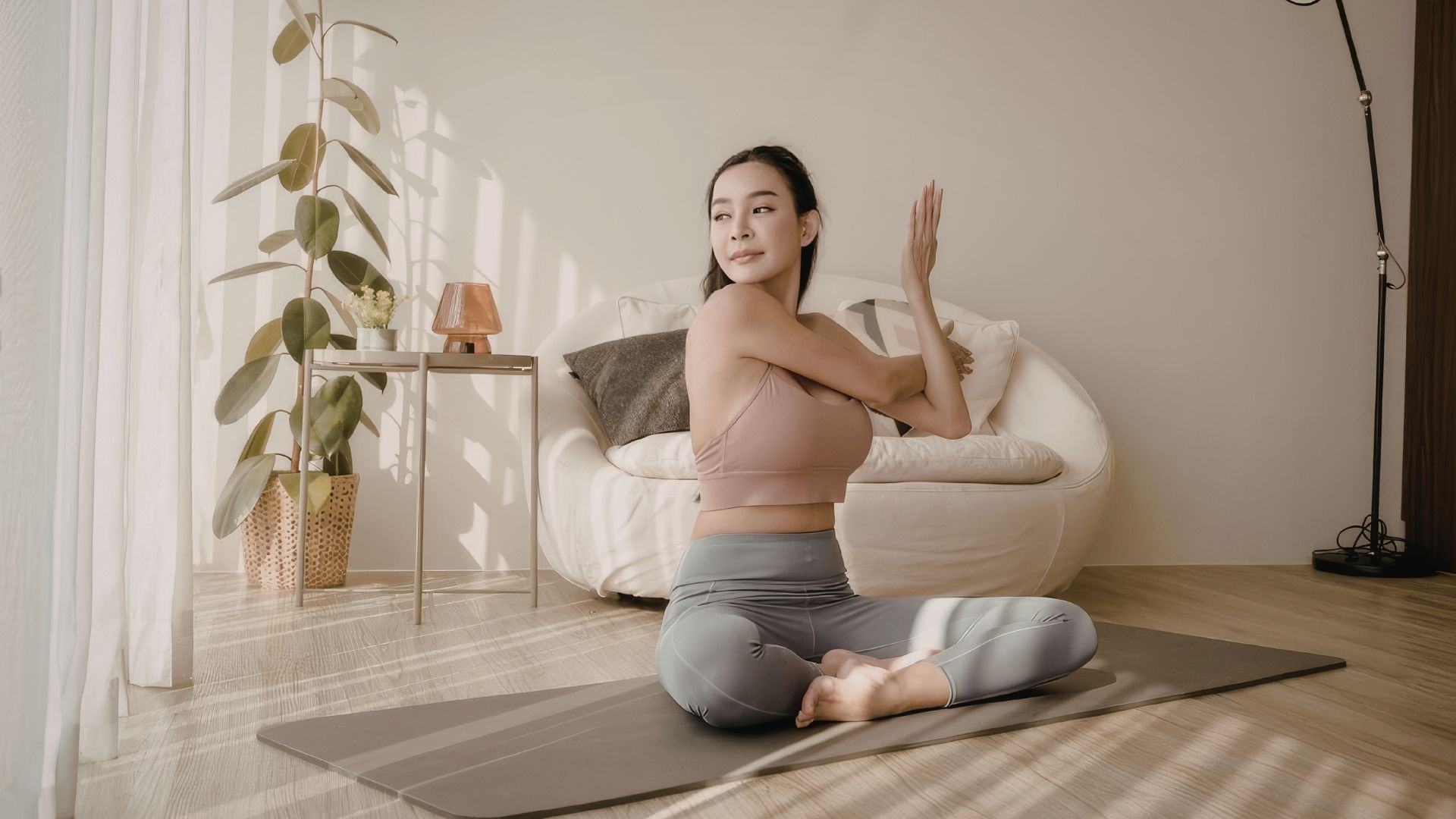 Mujer en postura sentada con las piernas cruzadas sobre una esterilla de yoga gris, interior luminoso con planta verde y sillón claro, que ilustra una práctica en el hogar