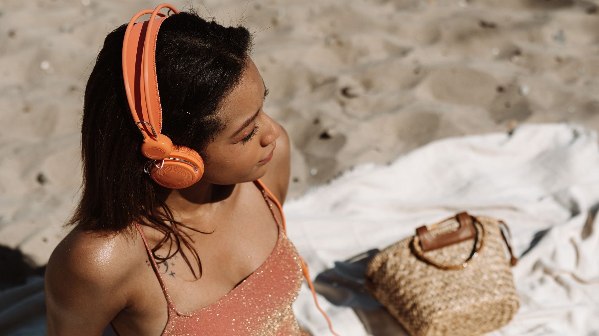 Eine Frau sitzt am Sandstrand mit einem orangenen On-Ear-Kopfhörer und einem Weidenkorb, die die Nutzung des kabellosen Kopfhörers im Urlaub und im Freien illustriert