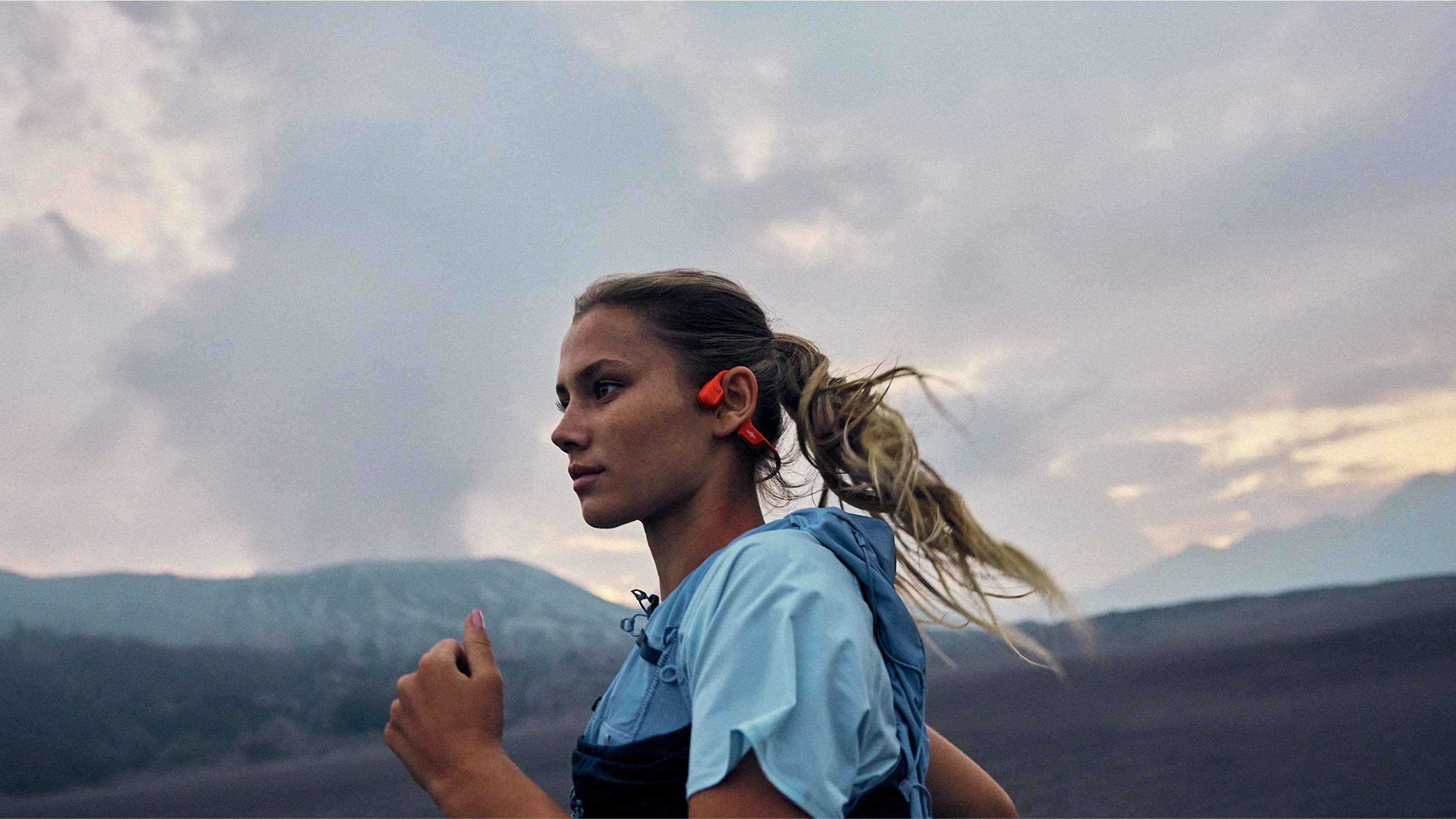 Coureuse en trail portant un casque à conduction osseuse orange avec sac d'hydratation, cheveux au vent, montagne et ciel nuageux au coucher du soleil en arrière-plan, illustrant l'usage du format en sport outdoor