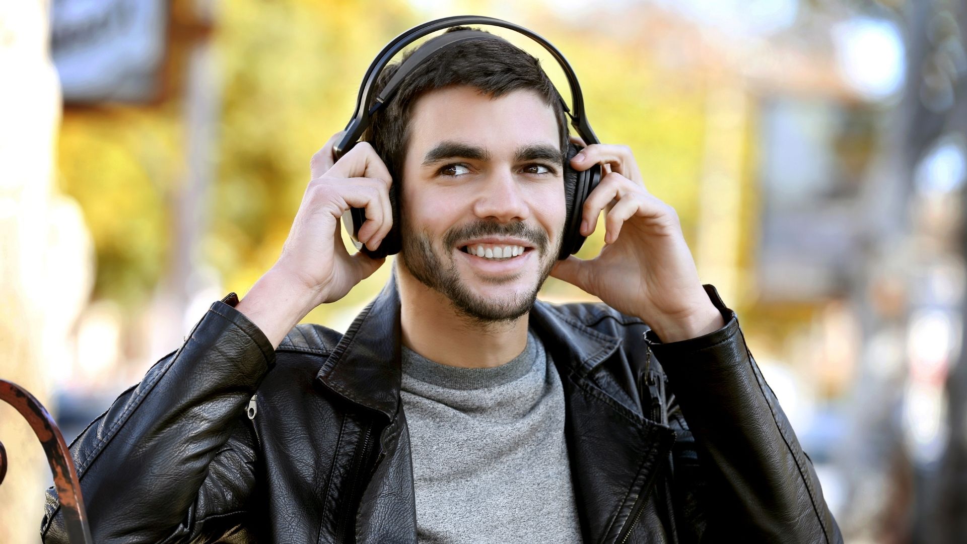 Homme souriant en blouson de cuir ajustant un casque circum-aural noir sur ses oreilles dans une rue urbaine ensoleillée, illustrant la transition bureau-extérieur et le travail mobile en tiers-lieu