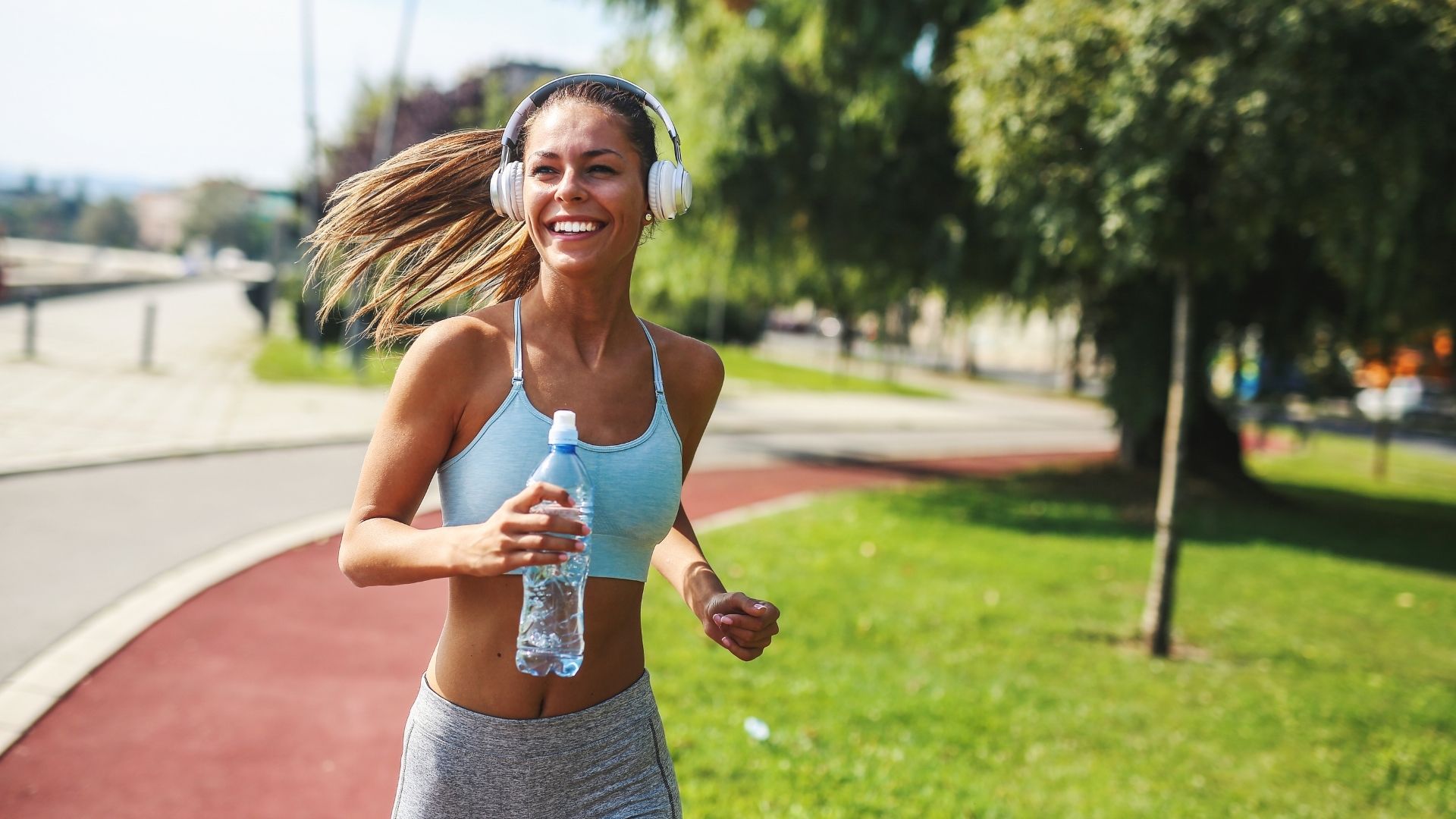 Coureuse souriante avec casque circum-aural blanc, bouteille d'eau à la main, sur piste rouge en milieu urbain ombragé, illustrant la course en environnement ouvert où la perception du trafic reste critique