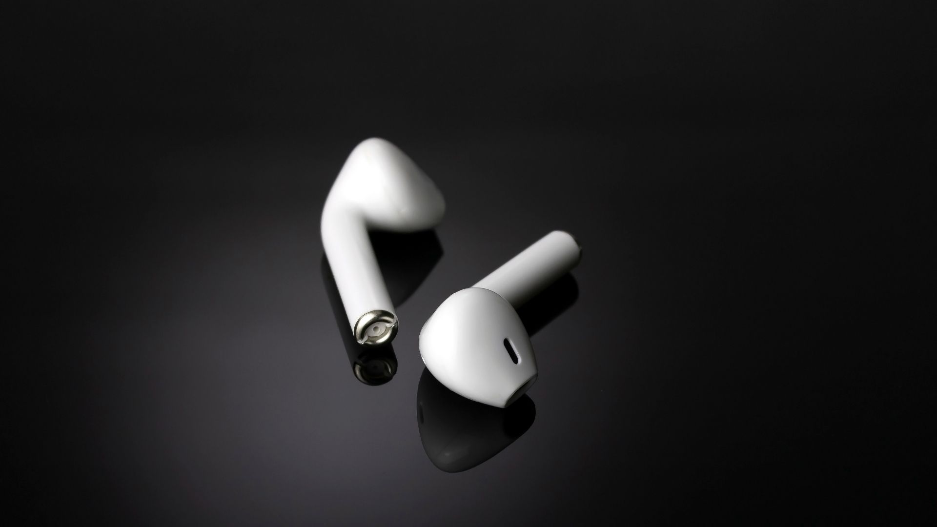 Paire d'écouteurs semi-in-ear blancs avec tige posés sur surface noire réfléchissante en plan studio macro, vue de trois quarts mettant en valeur la tige et la grille acoustique