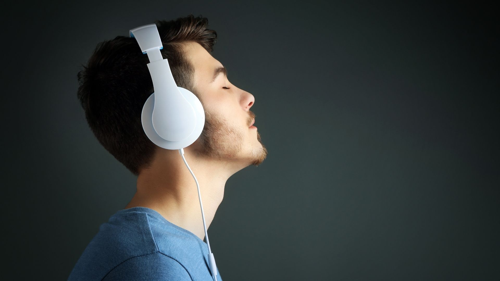 Jonge man in profiel met een witte bedrade circum-aurale hoofdtelefoon, ogen gesloten en hoofd licht achterover gekanteld, donkergrijze studio-achtergrond, illustrerend een geconcentreerde en attente luistersessie