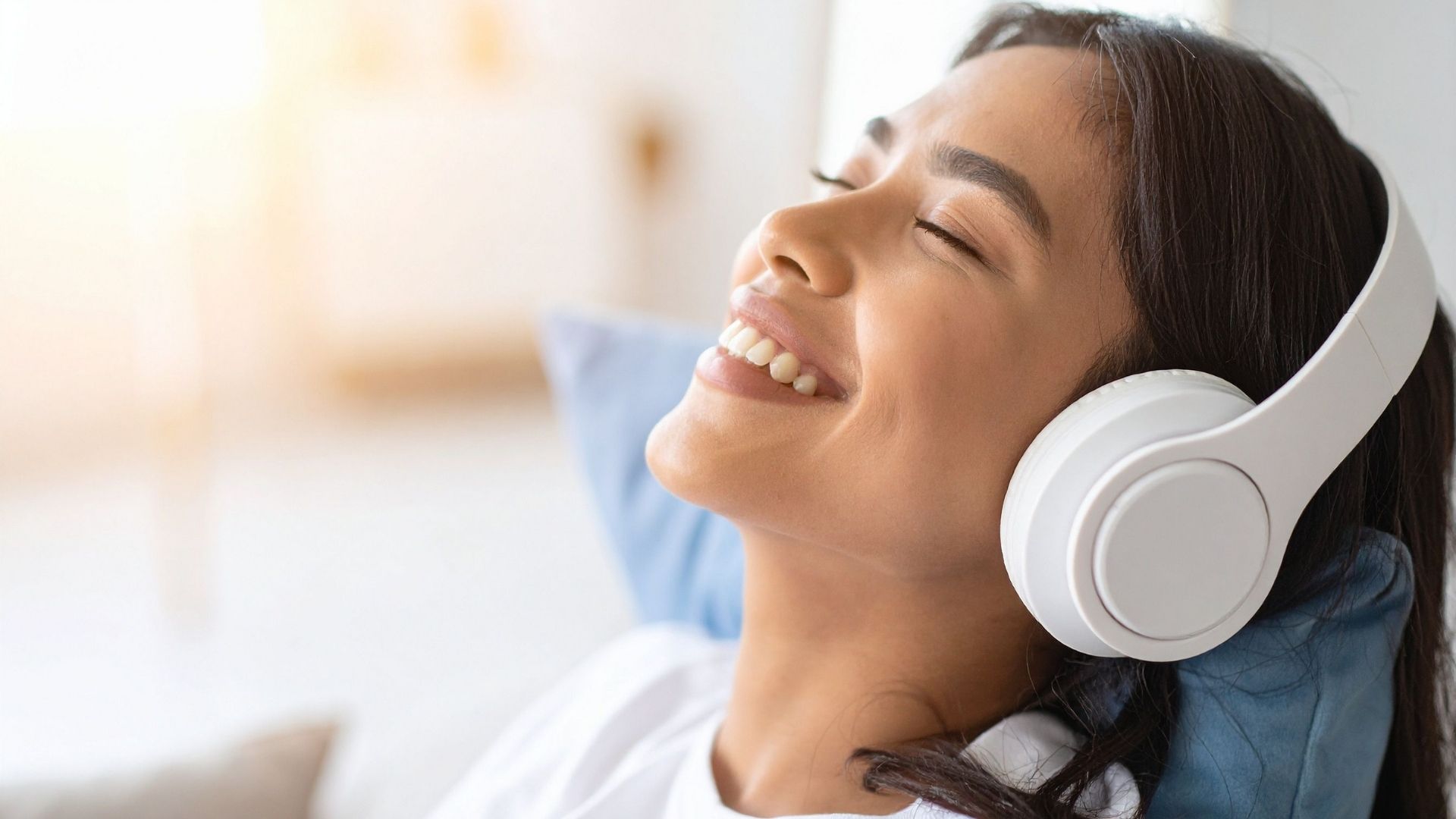 Vrouw glimlachend met gesloten ogen, liggend tegen een lichtblauw kussen, met een witte over-ear hoofdtelefoon, zacht natuurlijk licht in een lichte binnenruimte, die een ontspannen muziekbeluistering illustreert op een gecontroleerd volume